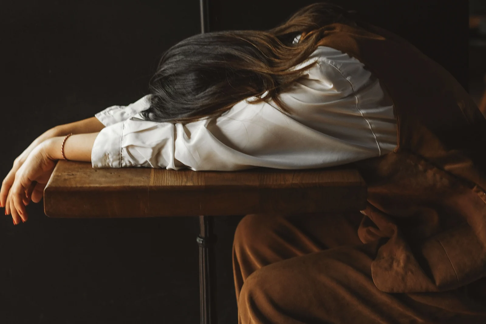 A woman slumped face-down on a small wooden table, exhausted, in low warm light