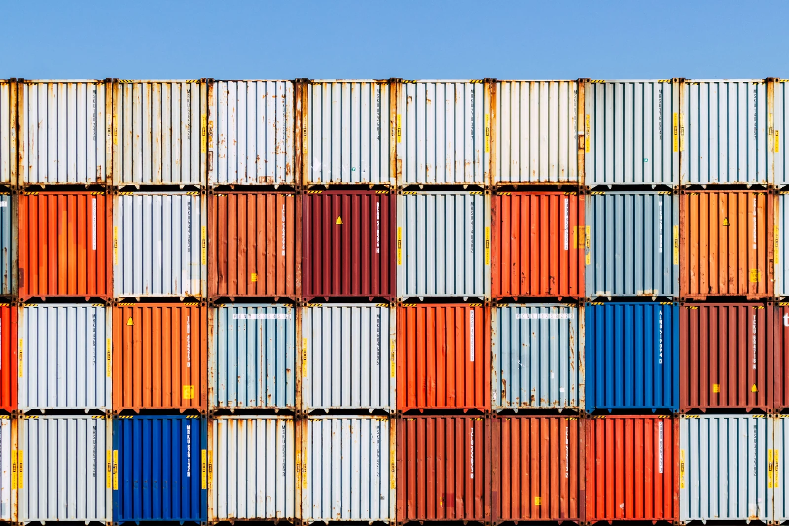 A wall of stacked shipping containers in mixed colors, most alike with a few standing out