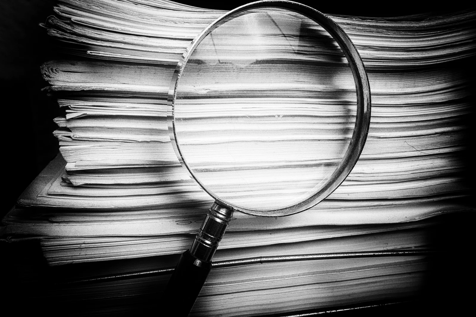 A magnifying glass resting on thick stacks of paper, high-contrast black and white