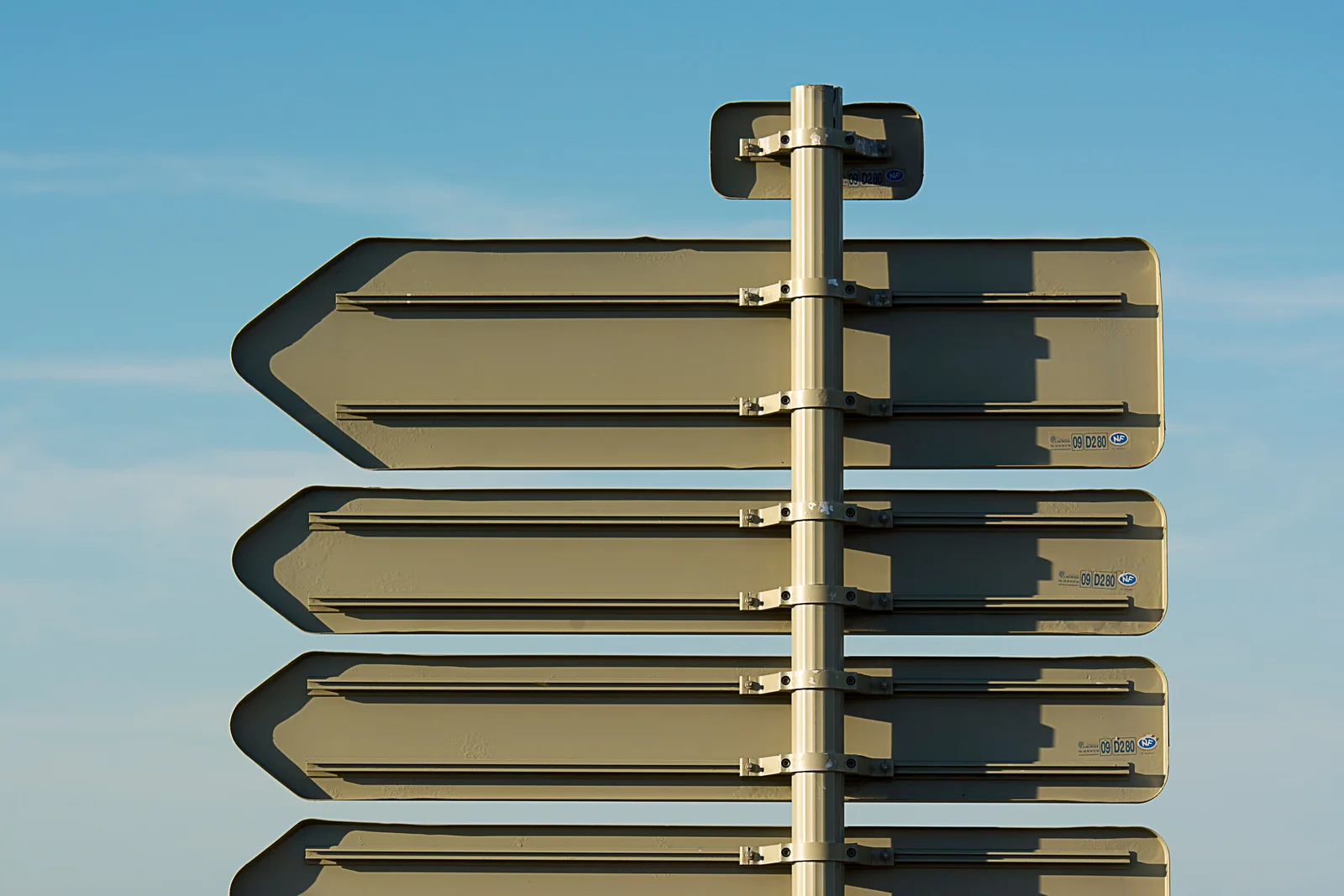 The backs of several blank directional road signs pointing different ways from a single pole against a clear blue sky