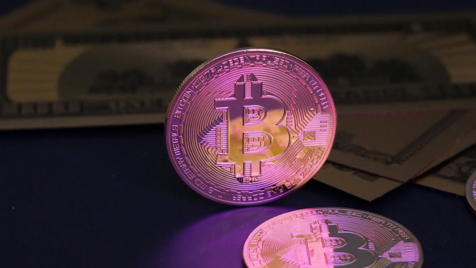 A Bitcoin coin lit in pink standing against hundred dollar bills in shadow