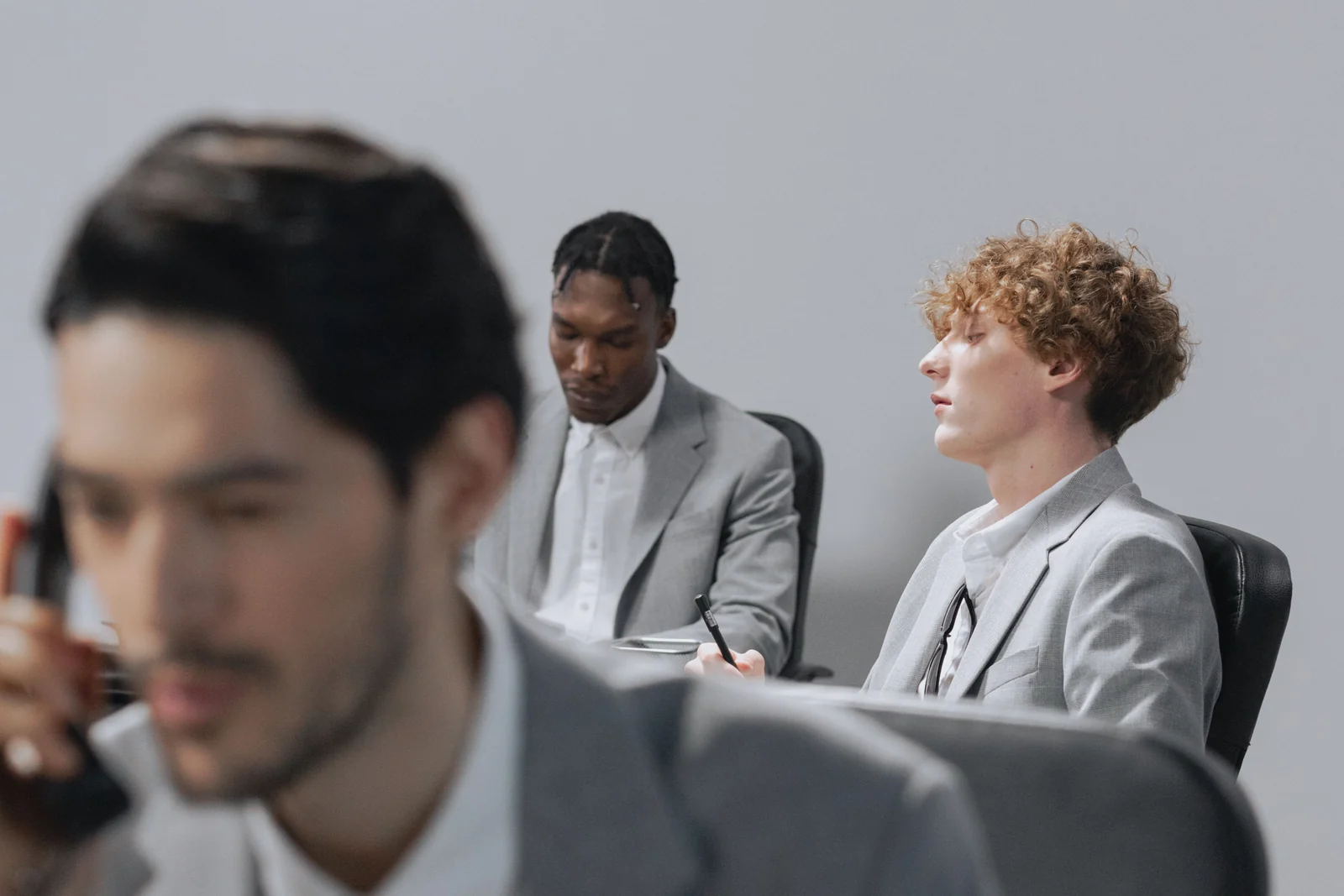 Three men in gray suits around a conference table, eyes closed and drifting mid-meeting