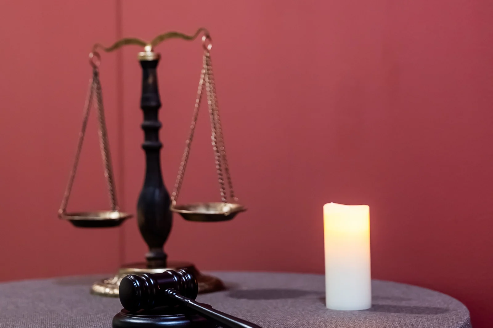 Brass scales of justice beside a gavel and a lit candle on a draped table against a red wall