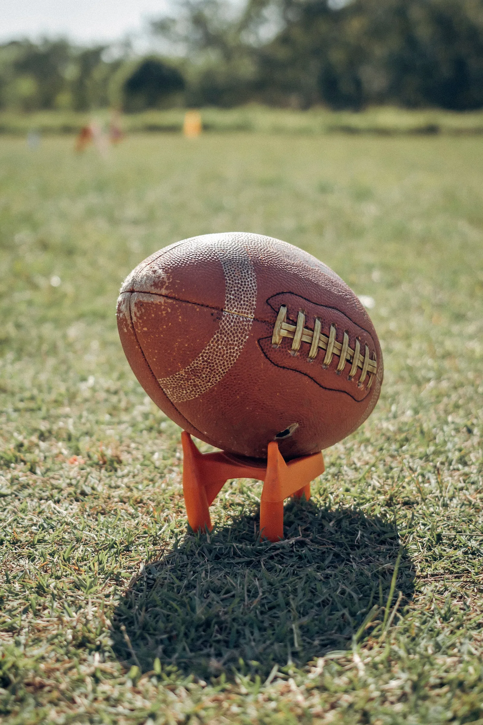 A worn football resting on an orange kicking tee in a sunlit field