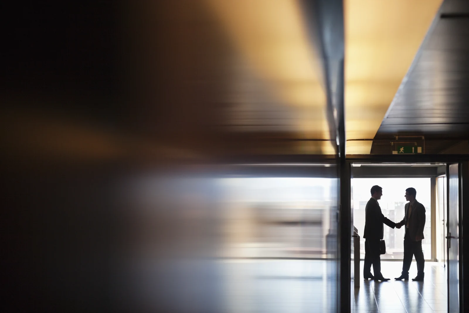 Two businessmen in silhouette shaking hands in a sunlit doorway at the end of a dark corridor