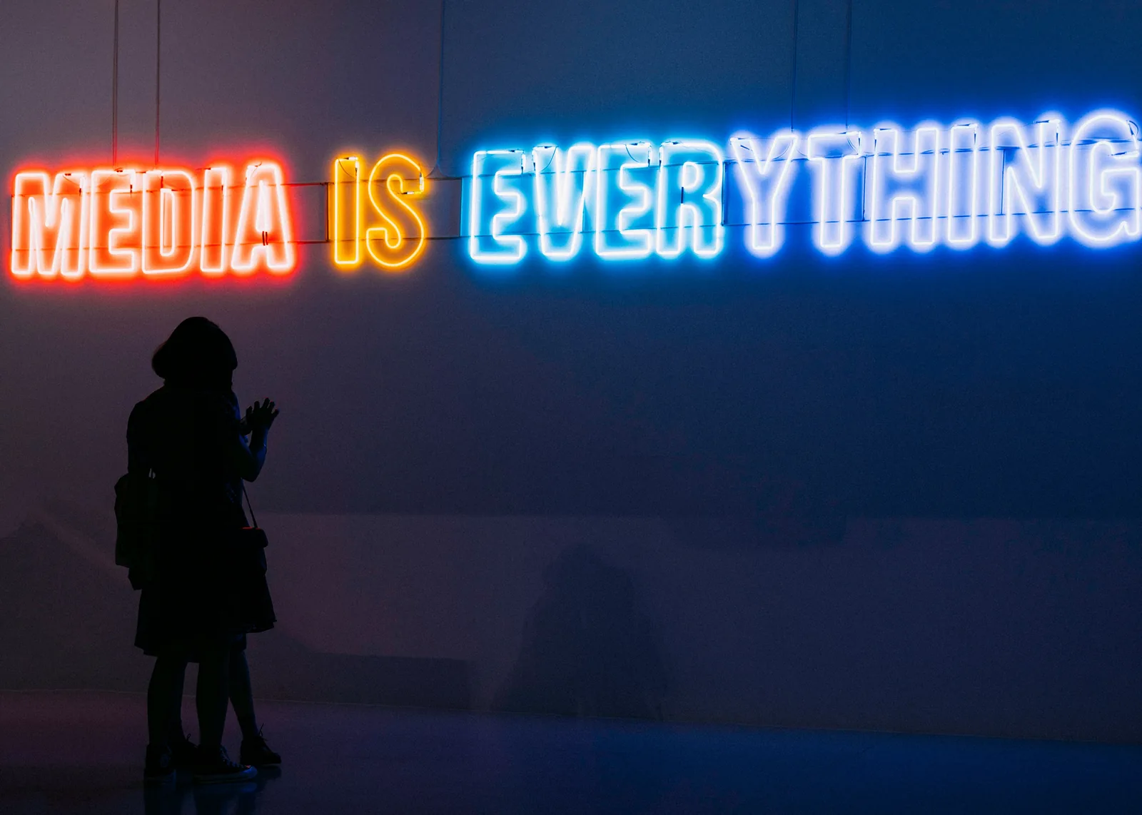 A silhouetted visitor facing a neon wall sign reading Media Is Everything
