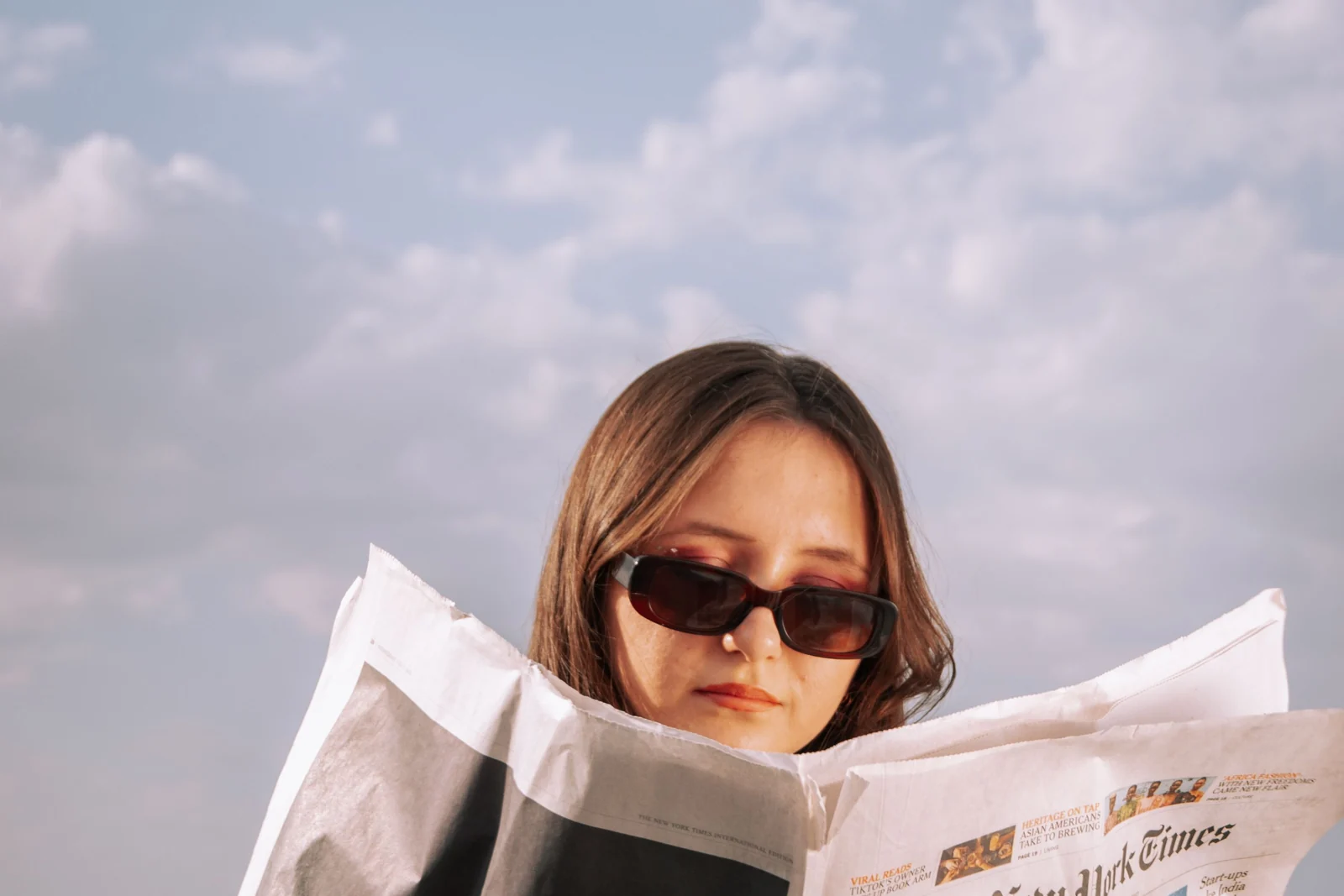 A woman in sunglasses reading the International New York Times outdoors under a wide sky