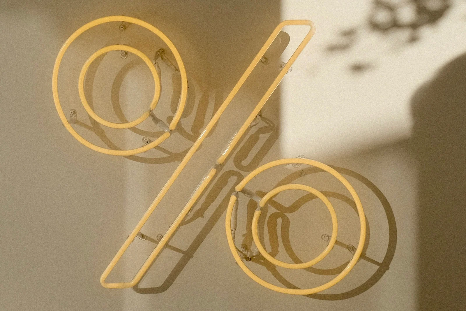 A neon percent sign in pale yellow casting a soft double shadow on a plaster wall