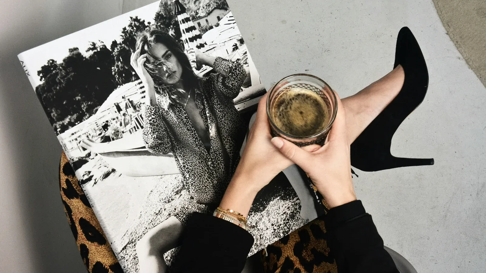 Hände halten ein Tumbler-Glas mit bernsteinfarbener Spirituose über einem Bildband, daneben ein schwarzer High Heel: Digitalmarketing und Werbung für Spirituosenmarken